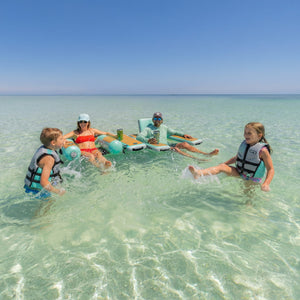 Family of four enjoying time in clear, shallow water with two kids with life vests on.