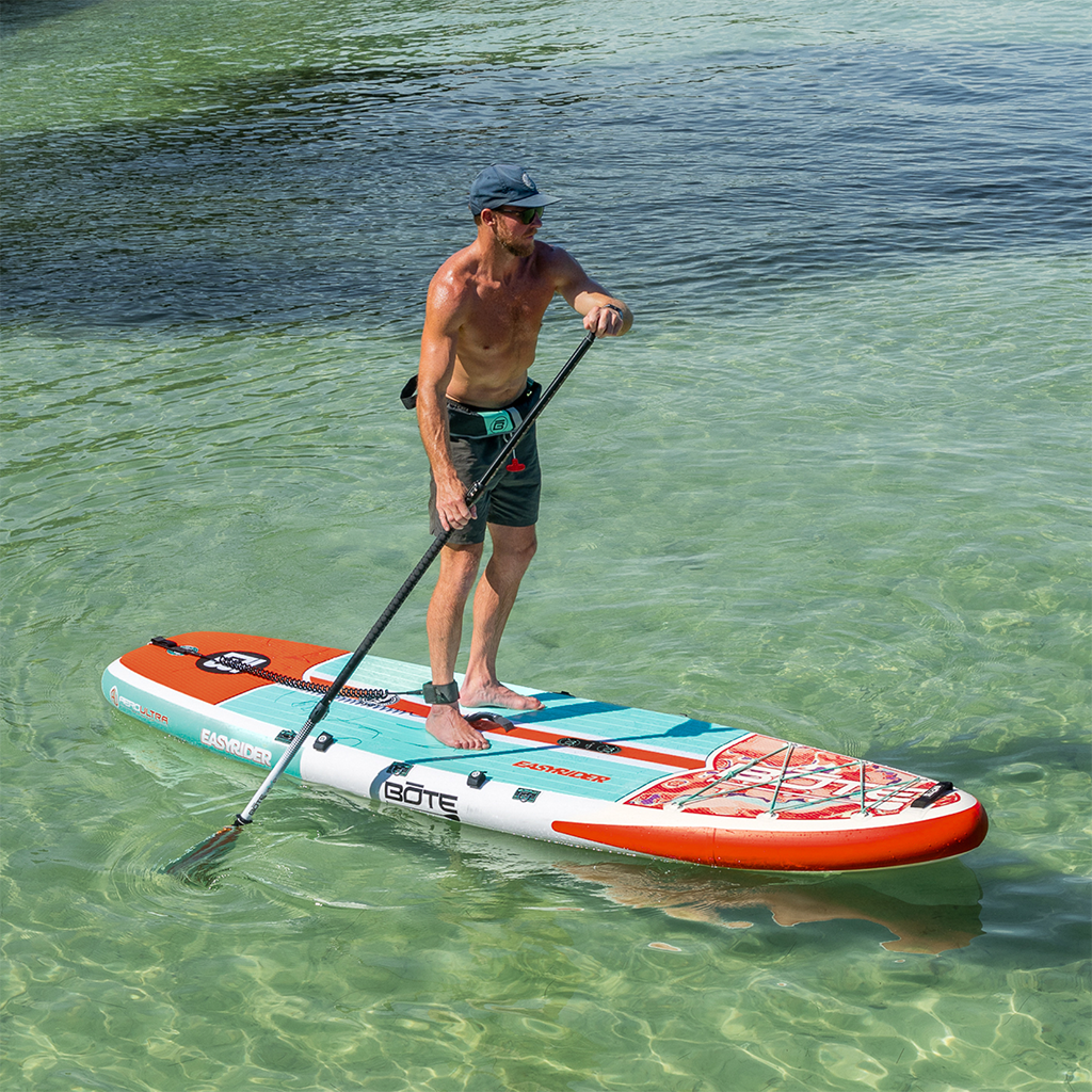A man stand up paddle boards on the EasyRider Aero in shallow water.