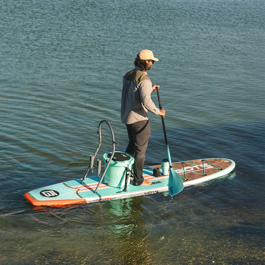 Man standing on a Breeze Gatorshell paddleboard with a cooler and RAC system on calm water.