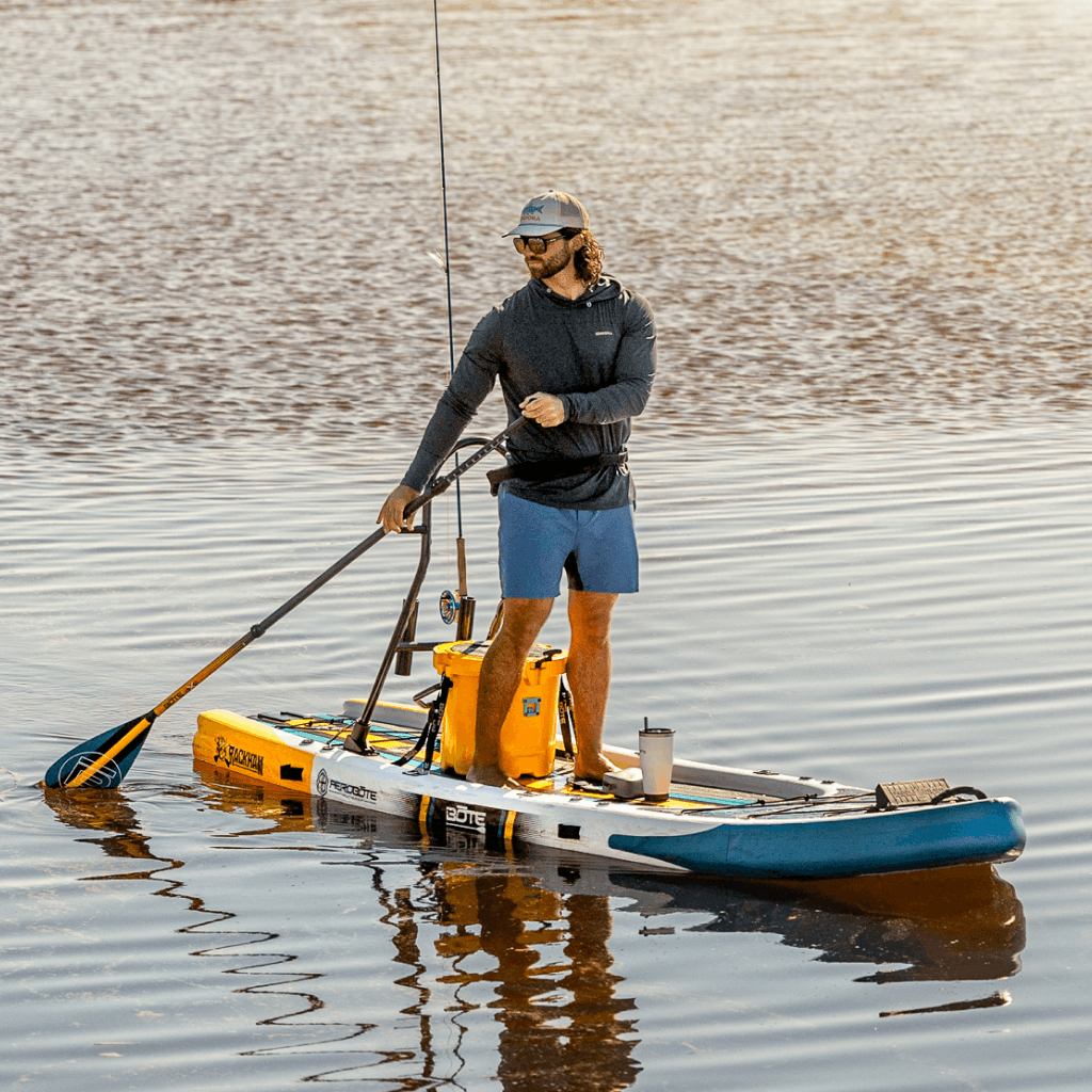 Angled view of Rackham Aero paddle board with cooler and fishing gear on calm water.