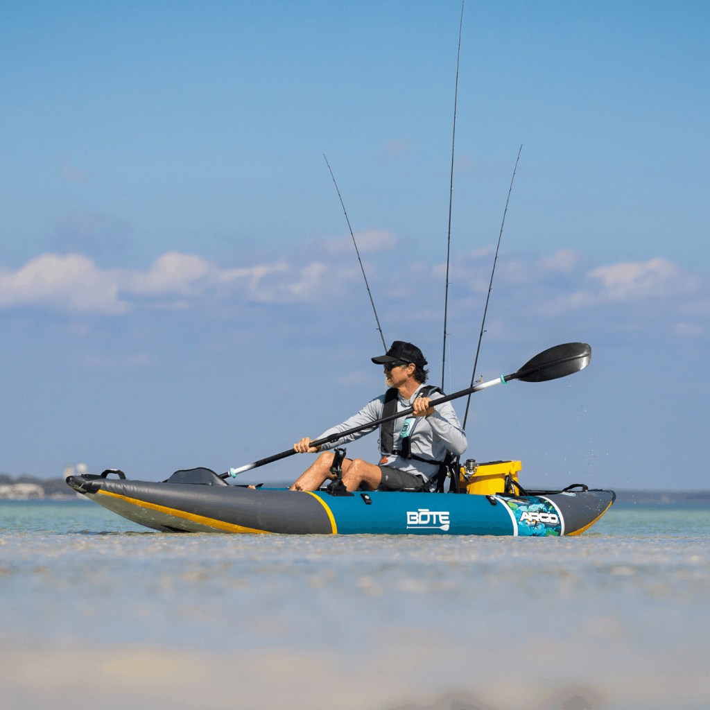 Man paddling a BOTE Argo inflatable kayak through shallow turquoise water with three fishing rods and a yellow cooler secured on deck.