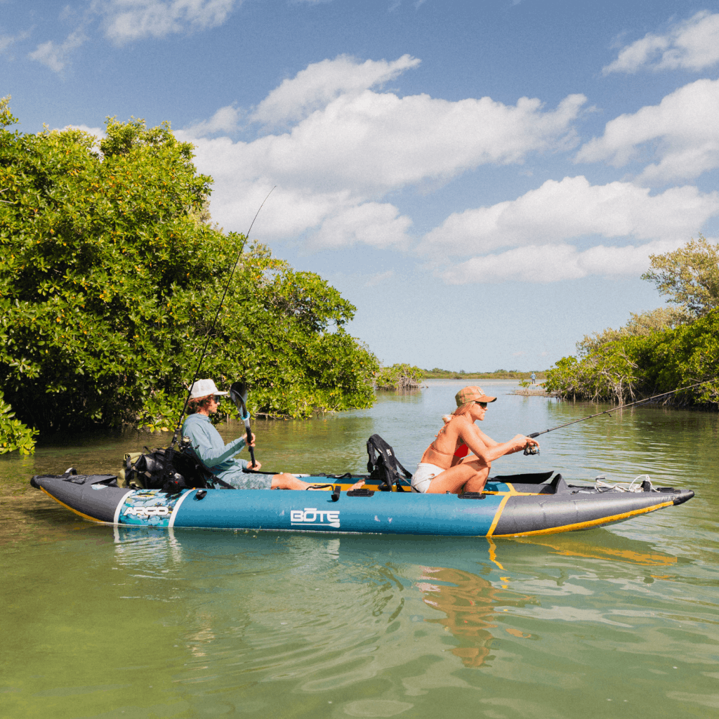 Two anglers fishing from a BOTE Argo teal and gray tandem inflatable kayak in a mangrove waterway, each holding a fishing rod.