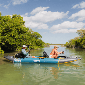 Two anglers fishing from a BOTE Argo teal and gray tandem inflatable kayak in a mangrove waterway, each holding a fishing rod.
