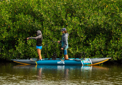 Two anglers standing and fishing from a BOTE Argo teal and gray inflatable kayak on calm water in front of a dense mangrove tree line.