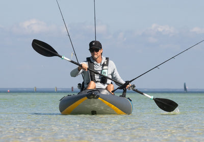 Front-facing view of an angler paddling a gray and yellow BOTE Argo inflatable fishing kayak in shallow coastal water with three fishing rods mounted on deck.