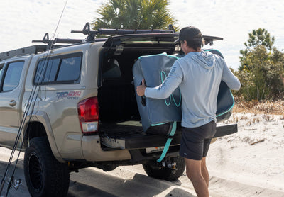 Person loading a gray BOTE Argo rolling travel bag into the bed of a truck on a sandy beach with fishing rods leaning against the vehicle.