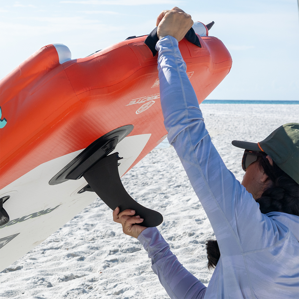 Woman installing a 10" Click-In Center Fin on an orange inflatable SUP on a beach.