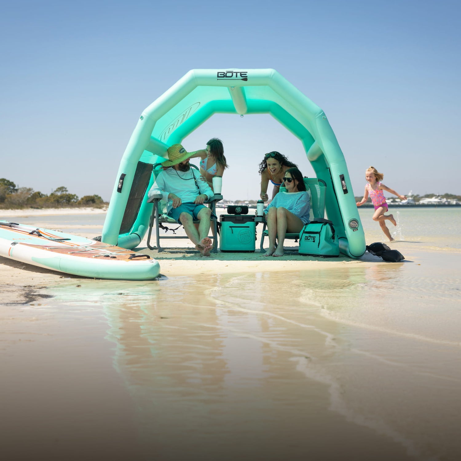 Family relaxing in chairs under the Hangout Shade inflatable beach shelter on a sunny sandbar.