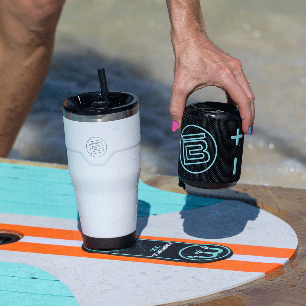 Hand placing a portable speaker next to a tumbler, both magnetically docked on a paddleboard.