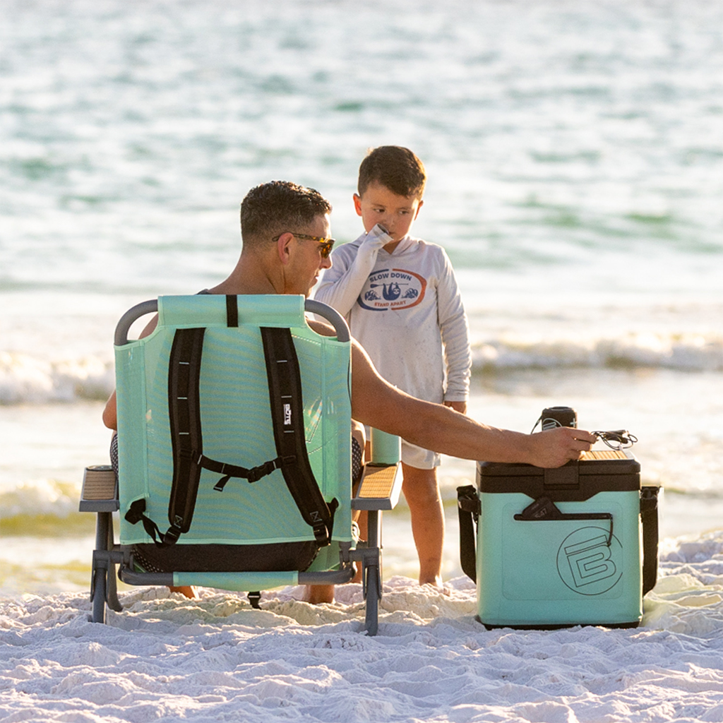 Rear view of person seated in Mule Dune Chair on beach with cooler and child nearby.