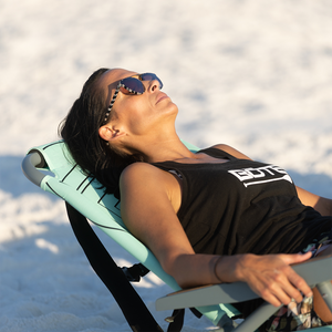 Person reclining in the Mule Dune Chair on sand wearing sunglasses and black tank.