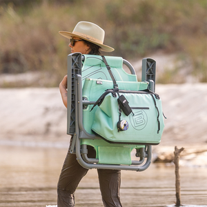 Rear view of person carrying folded Mule Outpost Chair with attached bag near water.