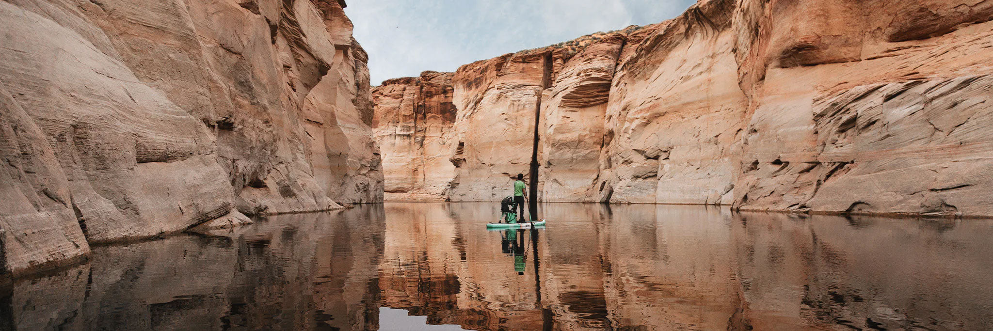 Paddleboarder with a dog glides through still water between towering red sandstone canyon walls.