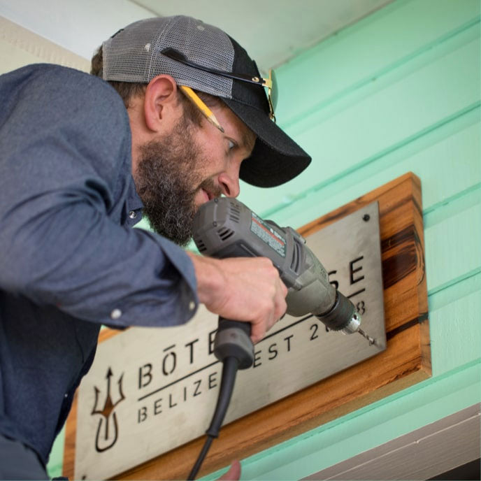 Man drilling screws into a wooden sign reading "BOTE House Belize" on a mint green wall.