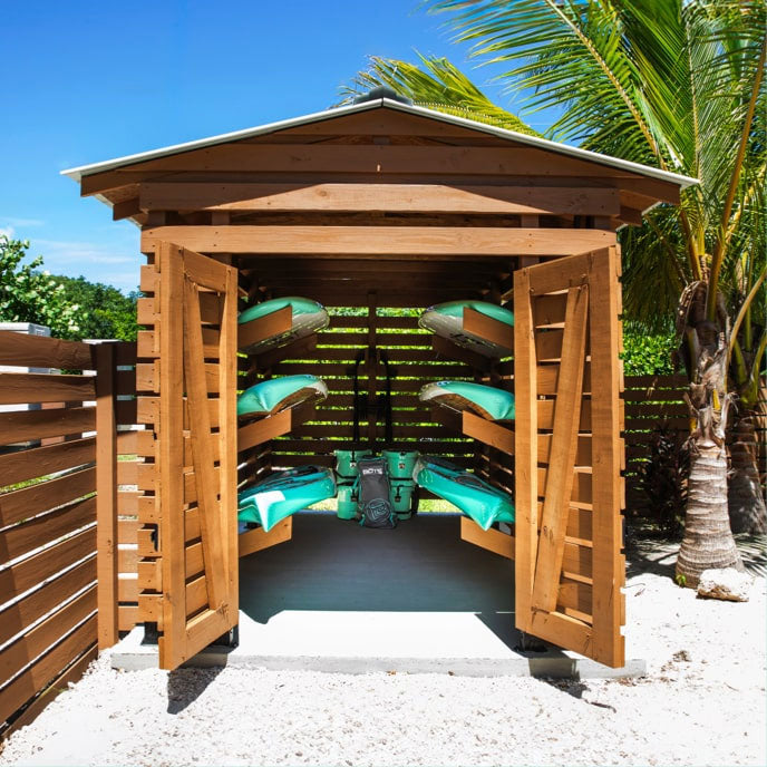 Open wooden storage shed on a sandy beach displaying stacked paddleboards and gear inside.