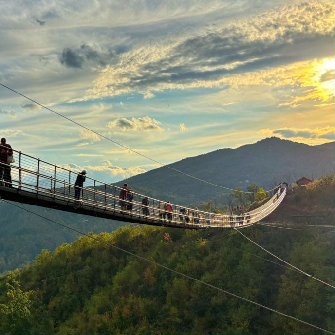 People walking across a suspended pedestrian bridge above a forested mountain valley at sunset.