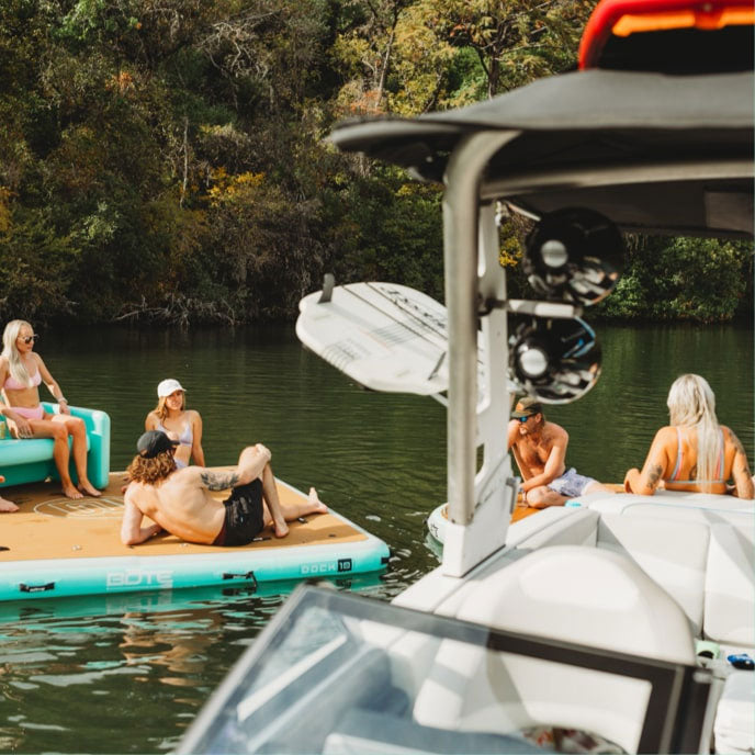 Group of friends relaxing on an inflatable dock platform next to a wake boat on a tree-lined lake.