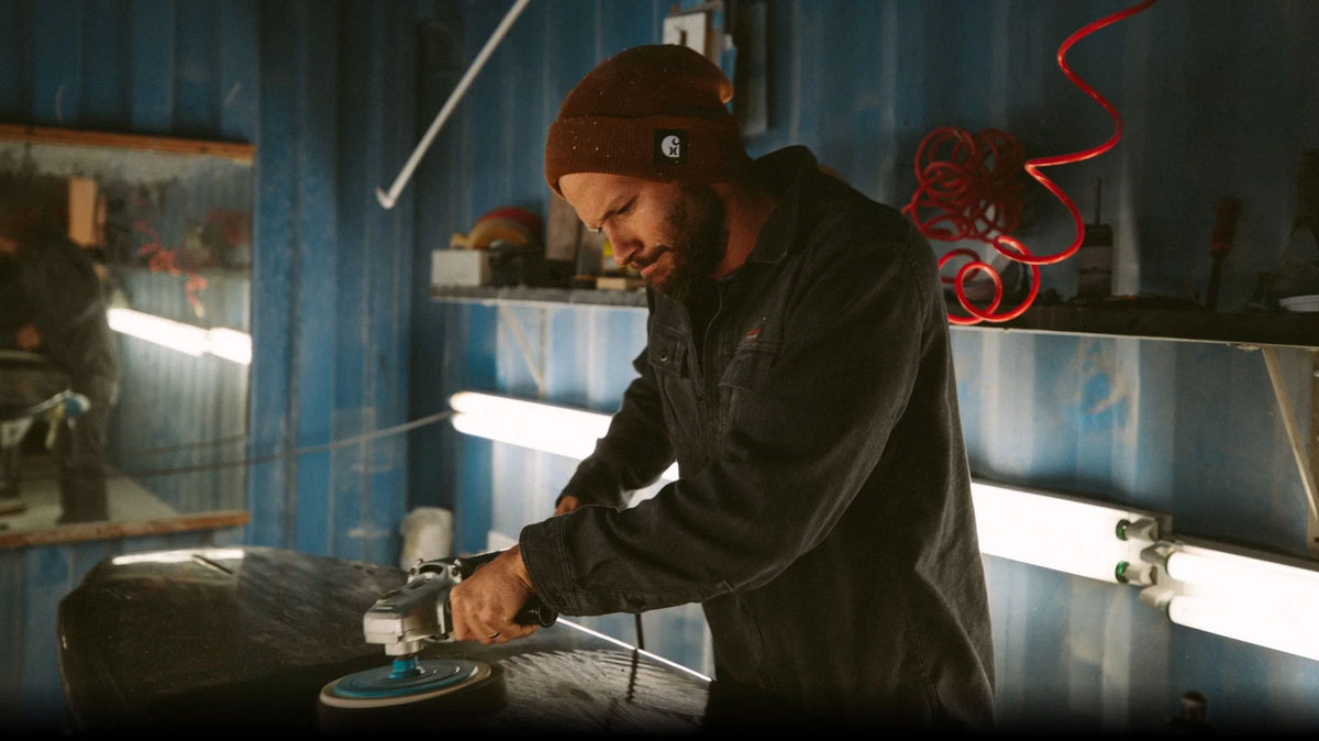 A man wearing a brown beanie and dark work jacket operates a grinder at a workbench inside a workshop with blue walls and bright fluorescent lights.