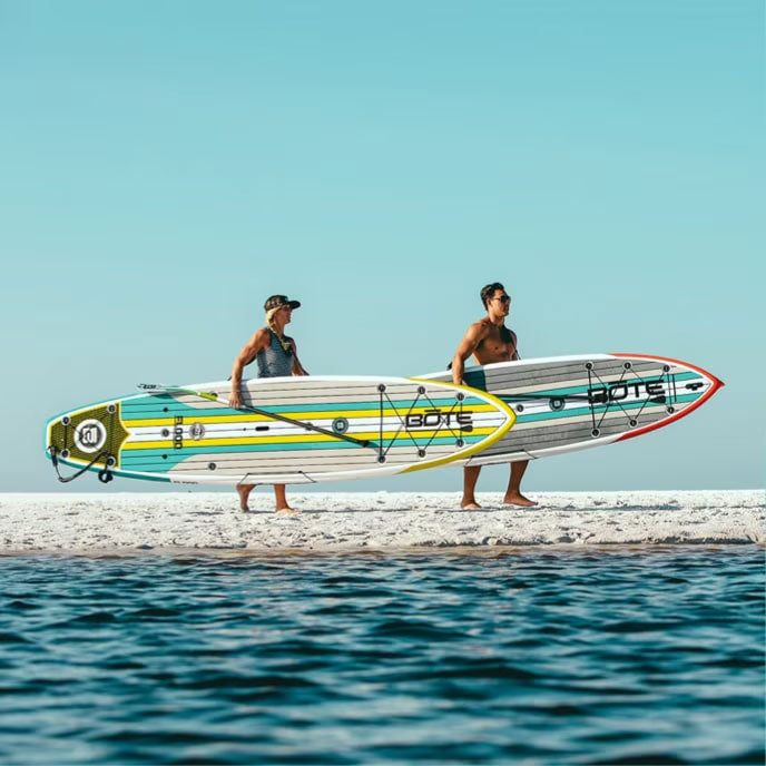 Two people carry paddleboards along a white sand beach under a clear blue sky.