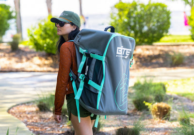 Woman carrying a gray and green inflatable paddleboard travel bag with BOTE branding walking on a path towards the beach.