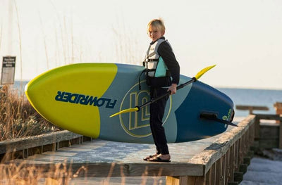 Kid standing on dock holding a Kids FlowRider Aero and paddle near the shoreline.