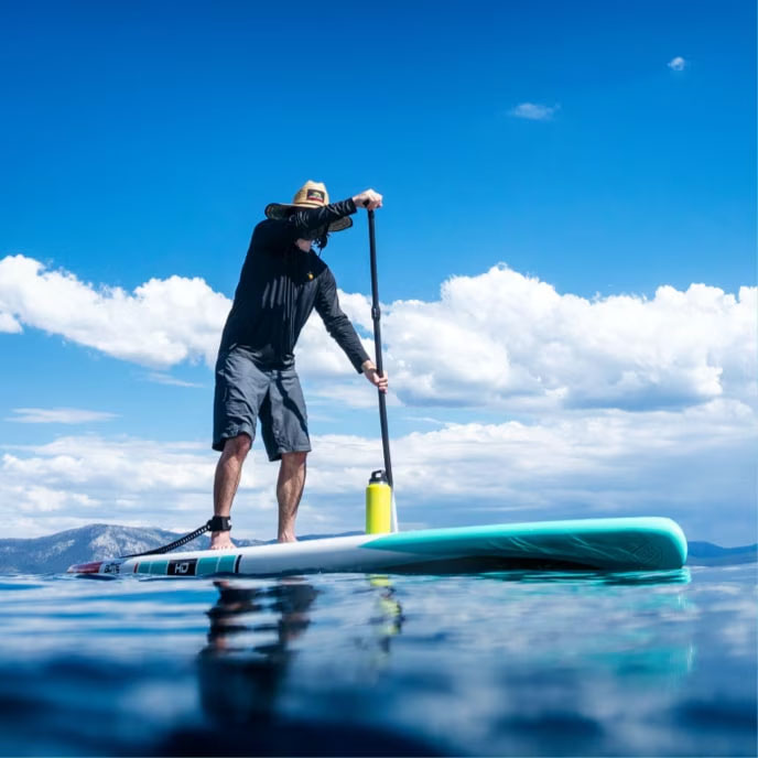 Man paddleboarding on calm blue water under a partly cloudy sky with mountains in the distance.