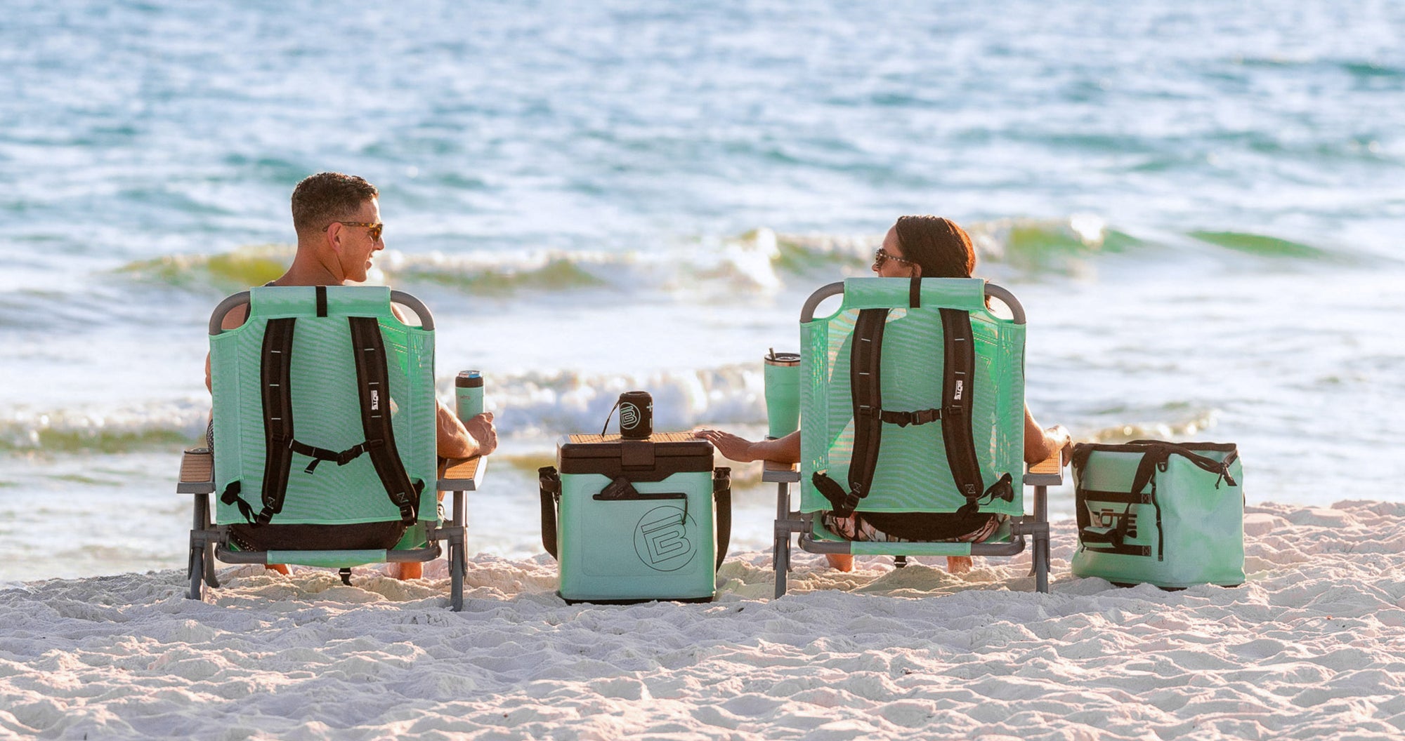 Rear view of two people seated in Mule Dune Chairs on beach with cooler between them.