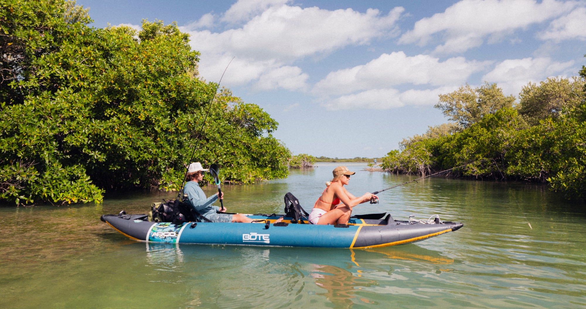 Two anglers fishing from a BOTE Argo teal and gray tandem inflatable kayak on calm green water surrounded by mangroves under a partly cloudy sky.
