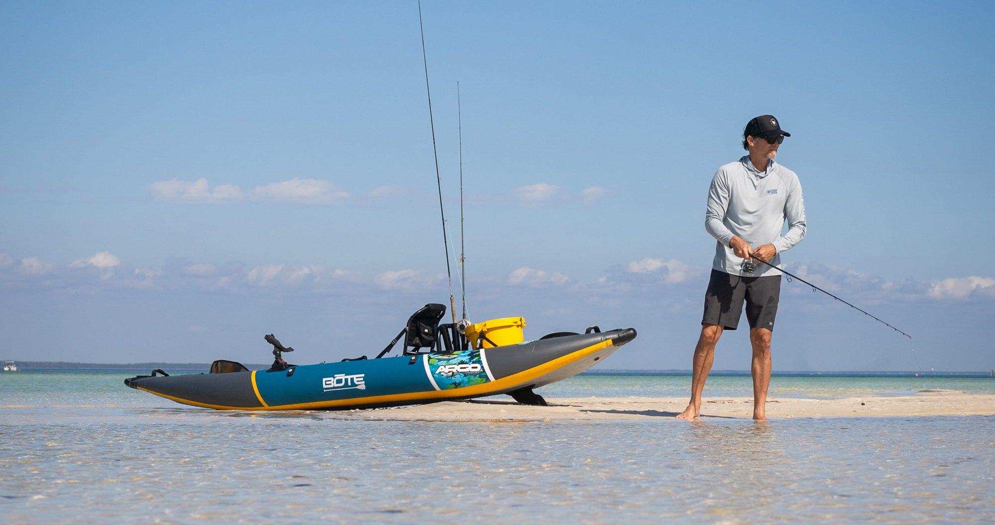 Angler standing on a shallow sandy shoreline holding a fishing rod next to a fully rigged teal and gray BOTE Argo inflatable kayak with a yellow cooler and fishing rods mounted on deck.