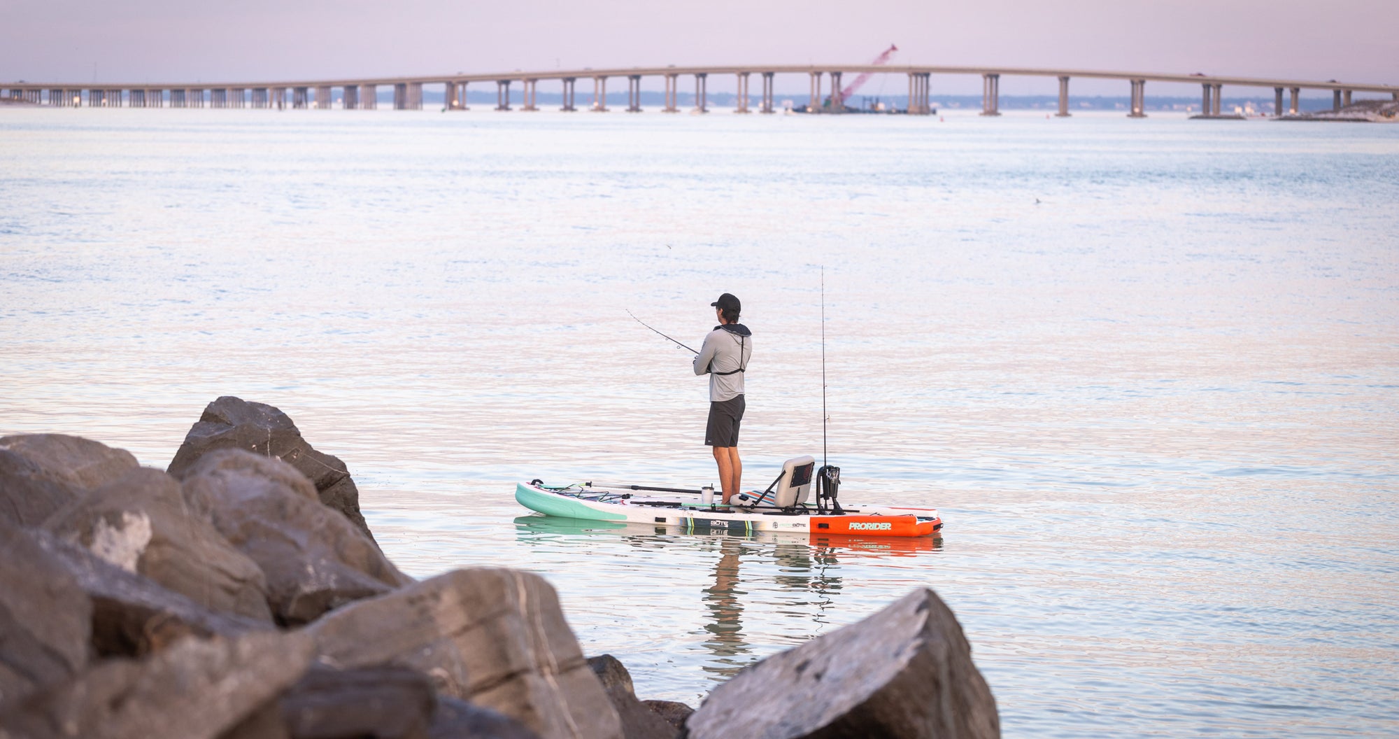 Person fishing from a stand-up paddleboard on a calm body of water with rocks and a bridge in the background.