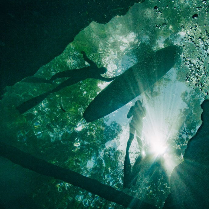 Underwater view looking up at silhouettes of a paddleboarder and swimmer seen through sunlit water.