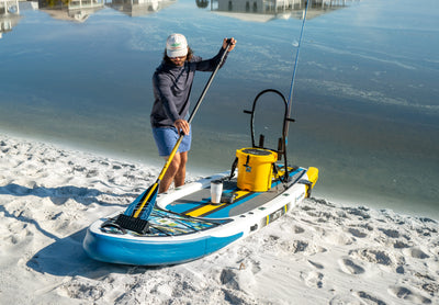 Angled view of Rackham Aero paddle board with cooler, paddle, and fishing gear on sand.