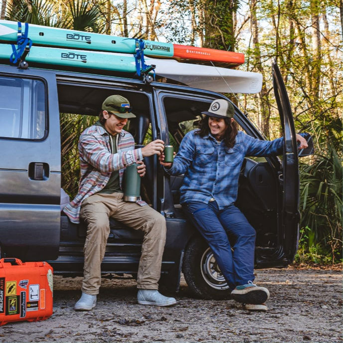 Two men sit in an open van with BOTE paddleboards on the roof rack, toasting with thermoses outdoors.