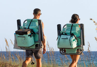 Rear view of two people walking to beach carrying chairs with attached cooler and gear bags.