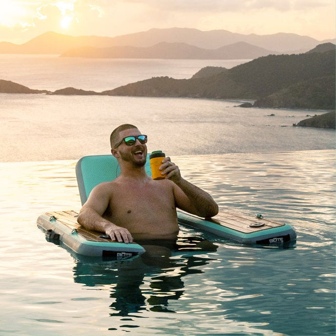 Man relaxing on an inflatable lounger in an infinity pool overlooking an island bay at sunset.