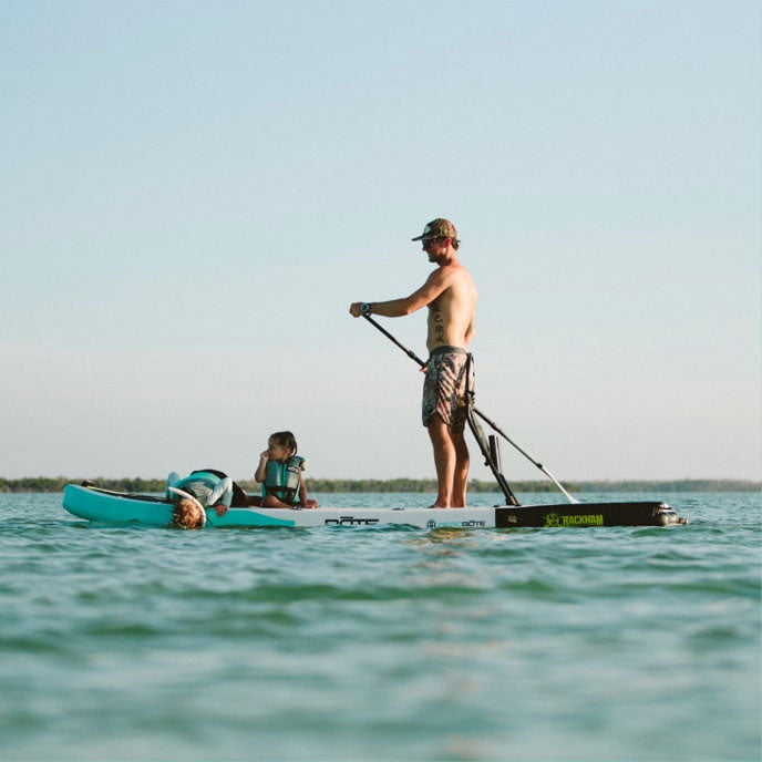 Man stands on a paddleboard on open water while two children play beside him.