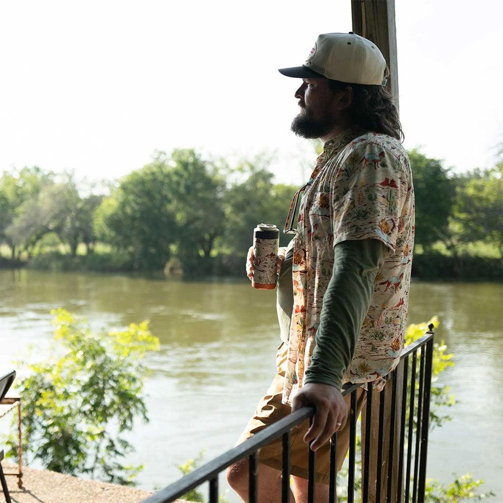 Man standing by a river, holding a coffee cup, with trees in the background